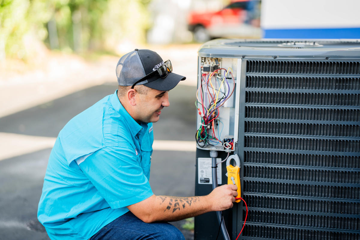 A Custom Air Conditioning & Air Quality technician performs AC service and repair by inspecting the wiring of an outdoor air conditioning unit with a yellow clamp meter