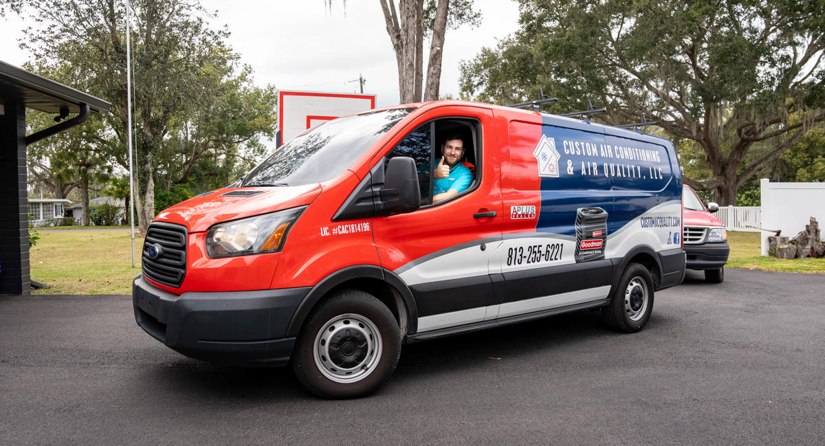 A Custom Air Conditioning Air Quality technician gives a thumbs up from the drivers seat of a branded company van showcasing the companys logo contact number and services 2