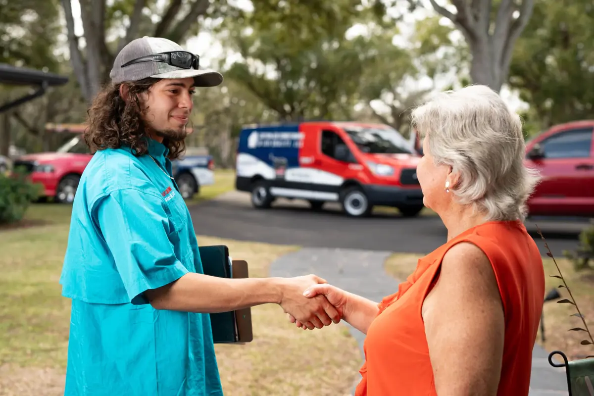 A Custom Air Conditioning Air Quality technician shakes hands with a customer outside her home with a branded company van visible in the background