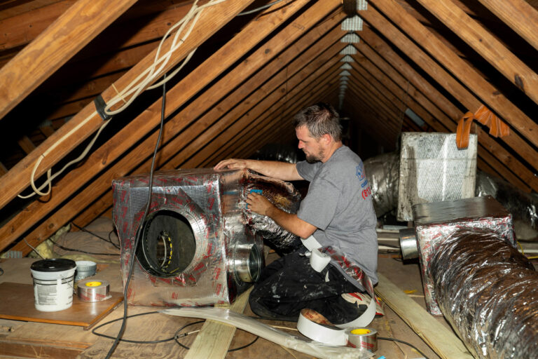 A technician works in a large attic sealing and insulating HVAC ductwork with metallic tape. The technician is kneeling next to a large insulated air handler 768x512 1