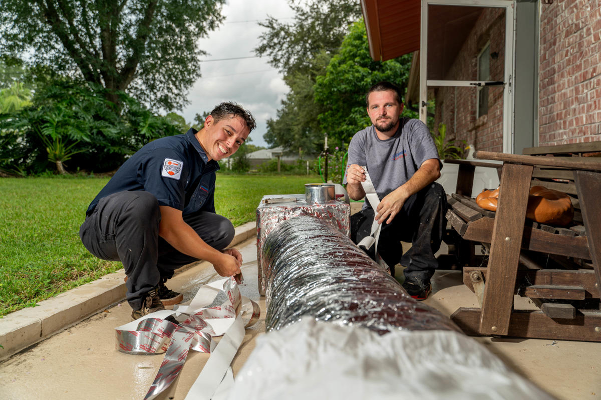 Two Custom Air Conditioning Air Quality technicians work on wrapping and sealing an insulated air duct outside a home both smiling and focused on the task 1