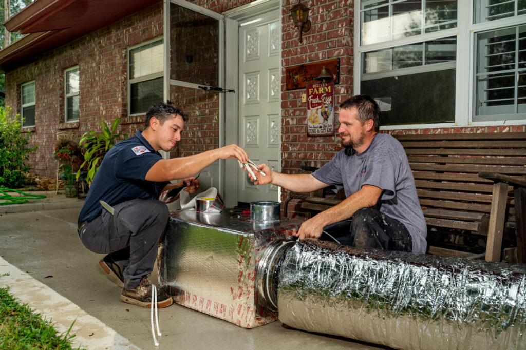Two HVAC technicians work together outdoors on a large insulated air duct. One technician hands a pair of pliers to the other as they collaborate on assembling ductwork 1024x682 1