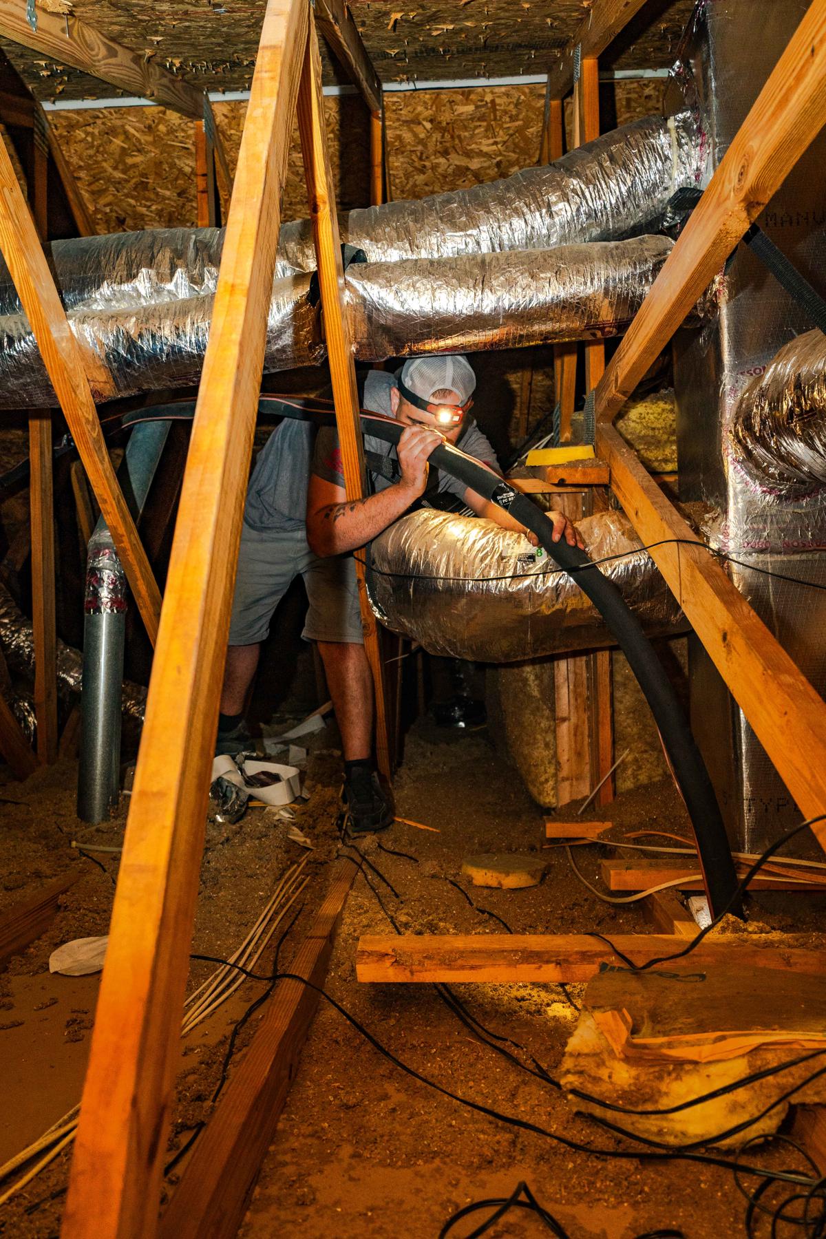 A technician in a tight attic space inspects and installs HVAC ductwork highlightinig the installation process for air conditioning systems by Custom Air Conditioning & Air Quality